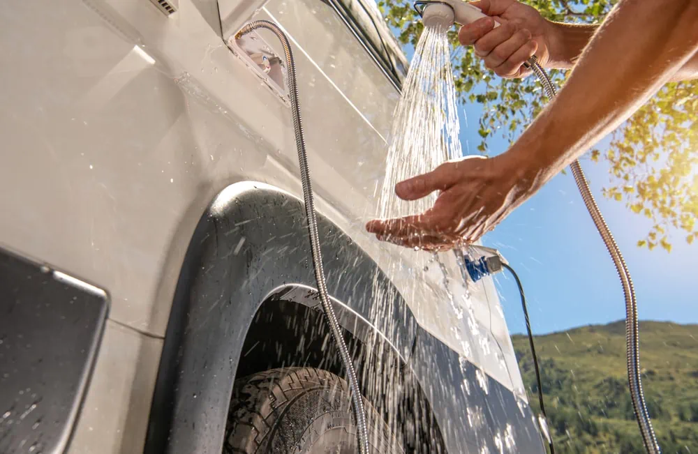 Hands using an outdoor shower attached to a white camper van. Water sprays over the hands against a backdrop of green hills under a clear sky.